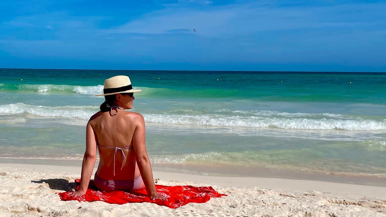 Beth looking over her right shoulder in bathing suit wearing a hat, sitting on an orange throw on the beach