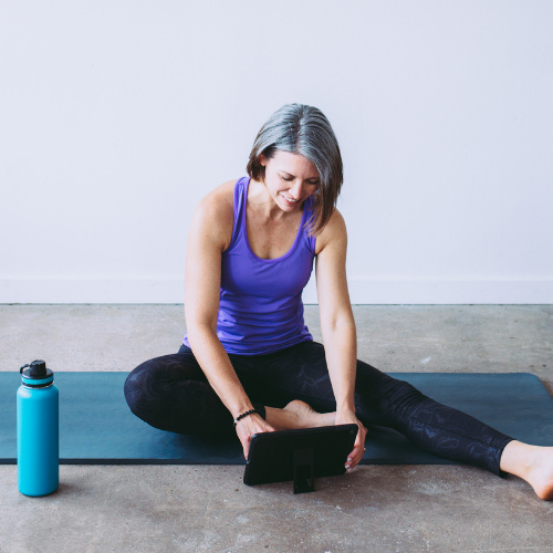 Beth on iPad sitting on gray mat. Wearing a purple top and black leggings with teal water bottle next to the mat.