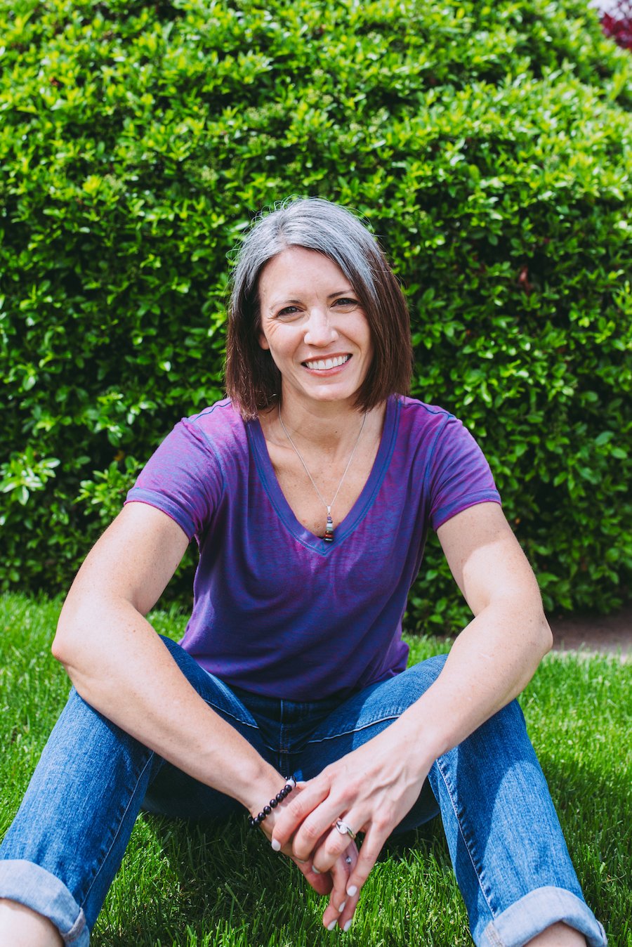 Beth smiling wearing purple top and blue jeans with her arms resting on her knees seated in the grass, green bush behind her.