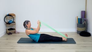 Beth in a teal top, black leggings, in Pilates roll up exercise, long band around her feet, hands holding band in a bicep curl position. On a gray Pilates mat in her home Pilates studio.