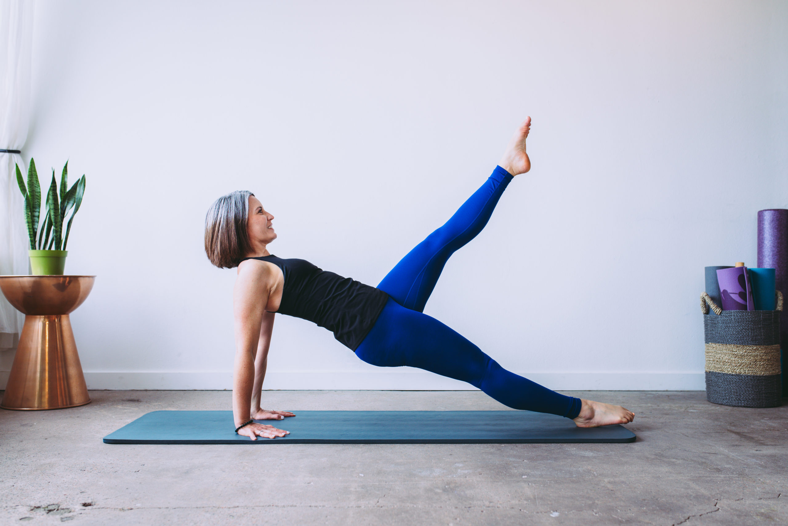 Beth doing Pilates mat exercise, balance control back wearing blue leggings and black top on a grey Pilates
