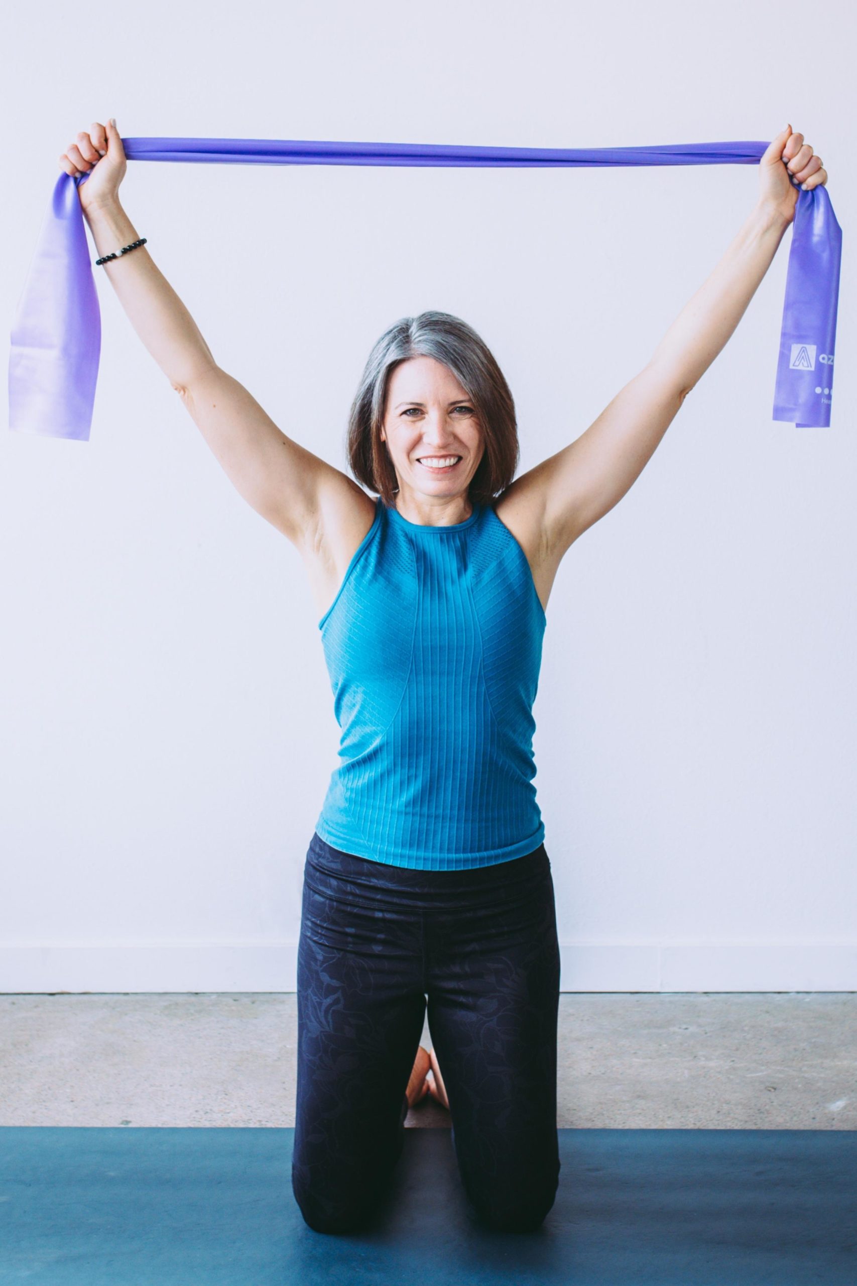 Beth kneeling on Pilates mat holding onto purple resistance band and reaching arms to the ceiling. Wearing teal top and black leggings. 