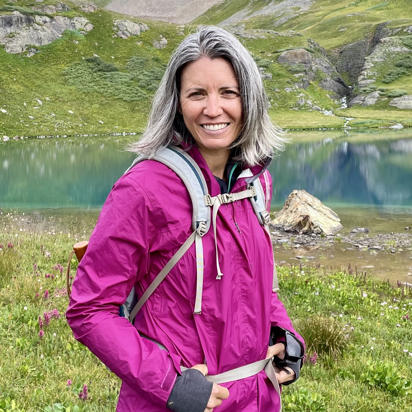 Beth wearing a magenta rain jacket and backpack in front of a Colorado lake