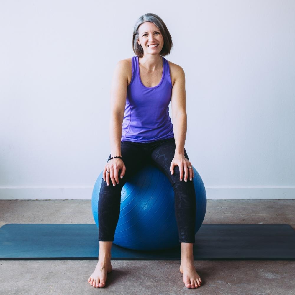 Beth smiling sitting on a large blue exercise ball wearing a purple top and black leggings.