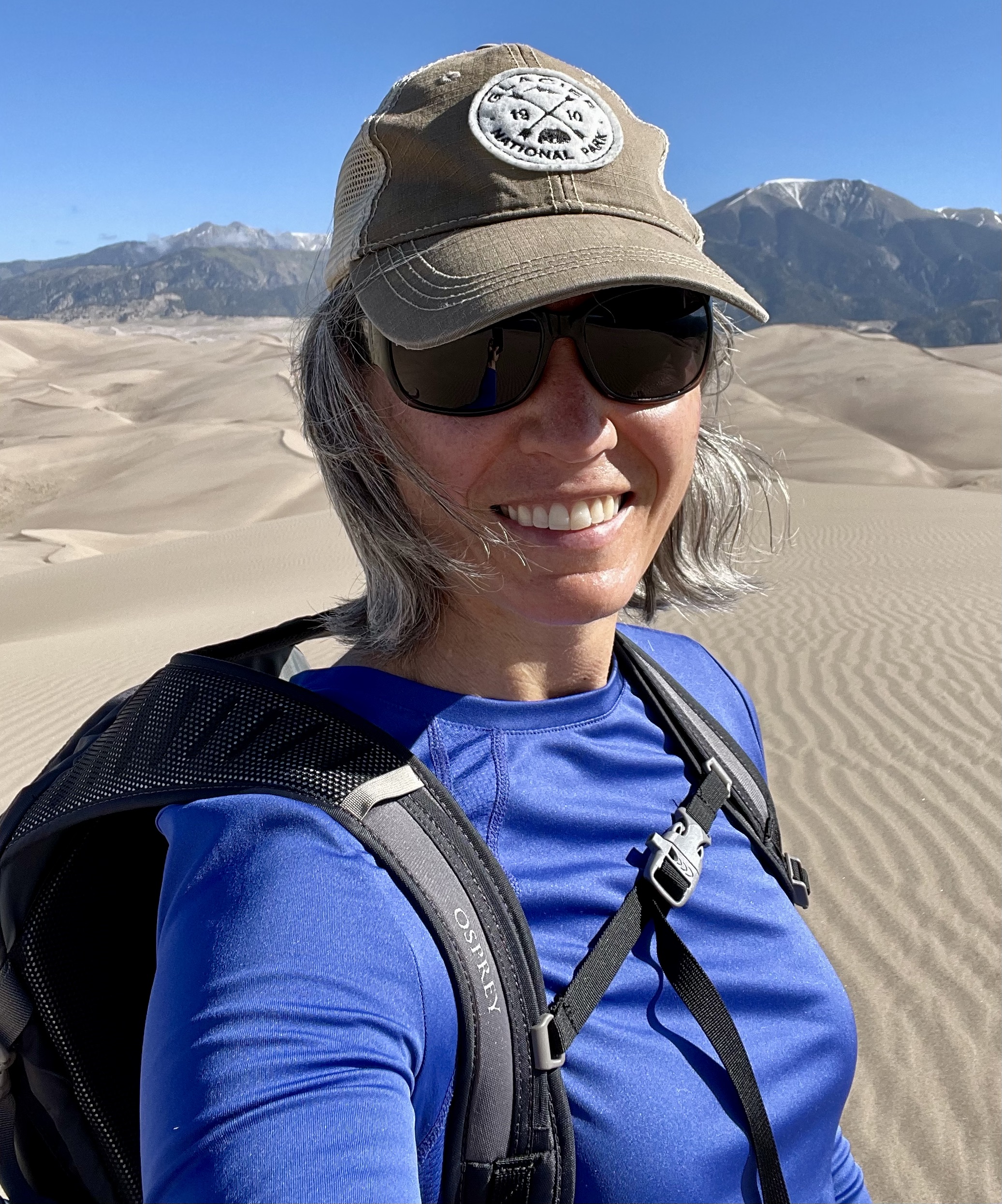 Beth in a blue shirt wearing a hat standing on top of Sand Dunes