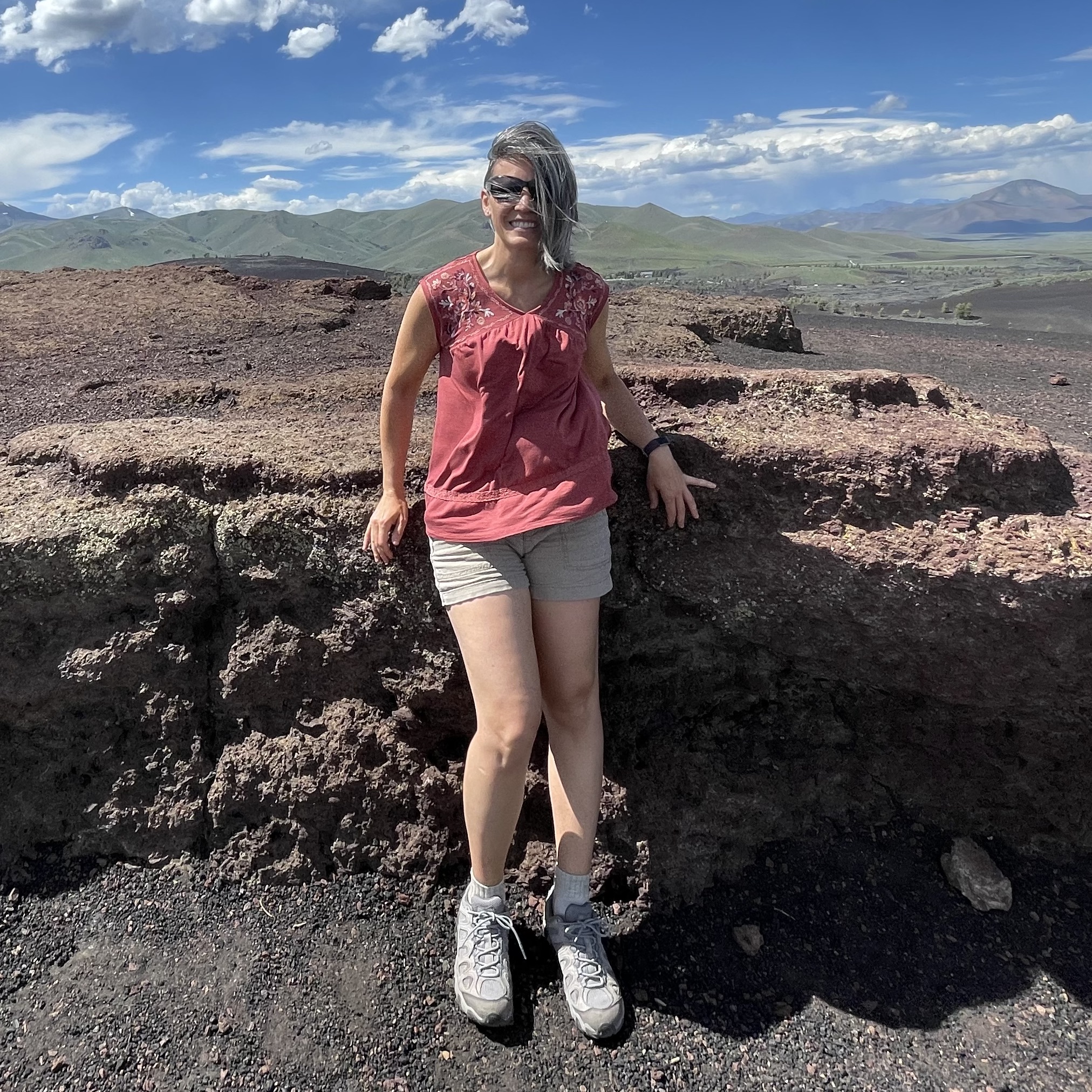 Beth in coral shirt and tan shorts standing in front of lava fields.